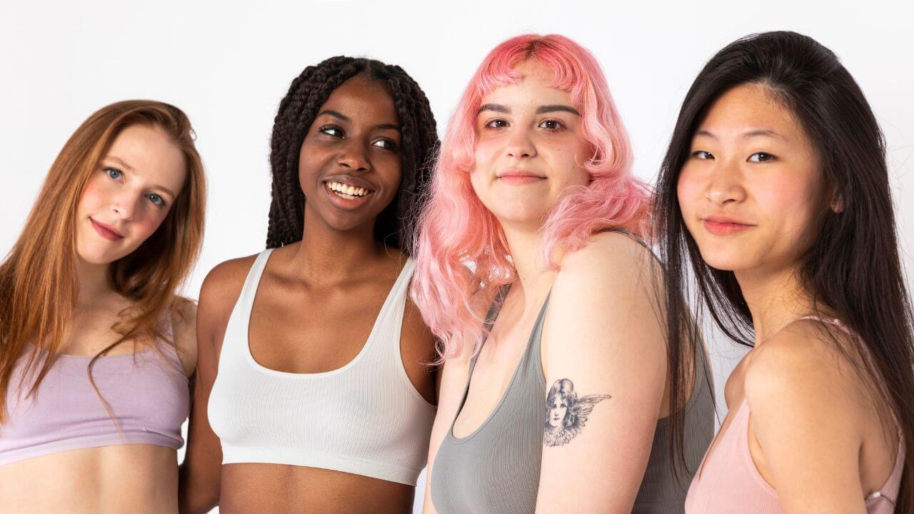 Quatro mulheres, a primeira ruiva, a segunda negra com tranças, terceira é uma branca com cabelo rosa e a ultima morena.