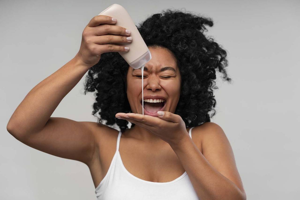 imagem de uma modelo de cabelos cacheados médios, apertando um shampoo com uma mão sob a outra mão, usando regata