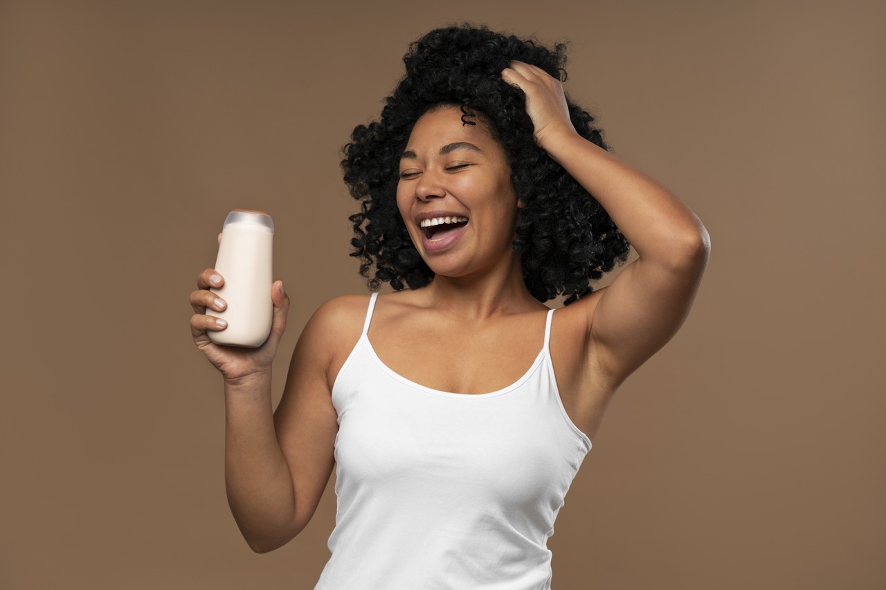 Imagem de uma modelo de cabelos cacheados médios, segurando um shampoo em uma mão e a outra no cabelo, de regata branca no fundo marrom