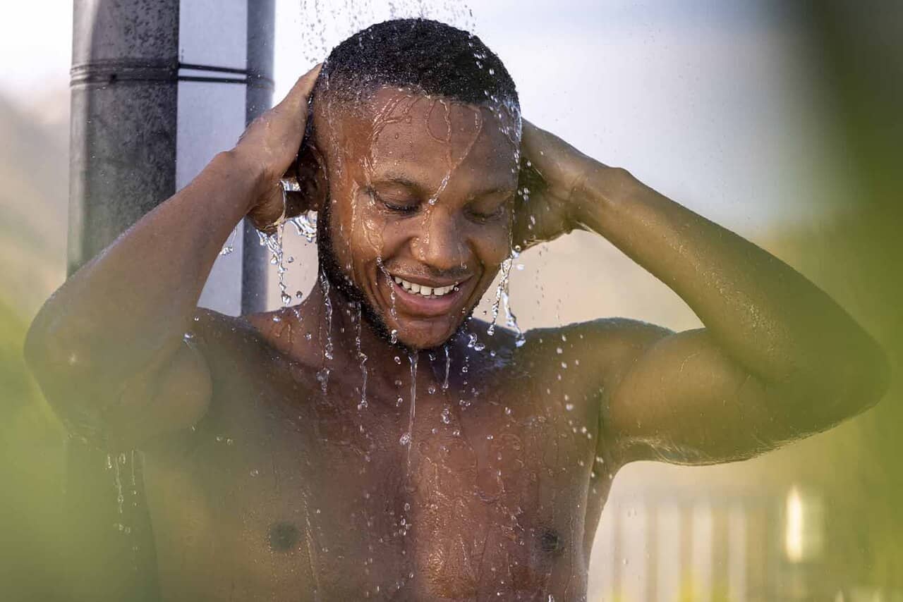 Imagem de um modelo de cabelo cacheado curto, tomando uma ducha com as mãos na cabeça, com o fundo desfocado