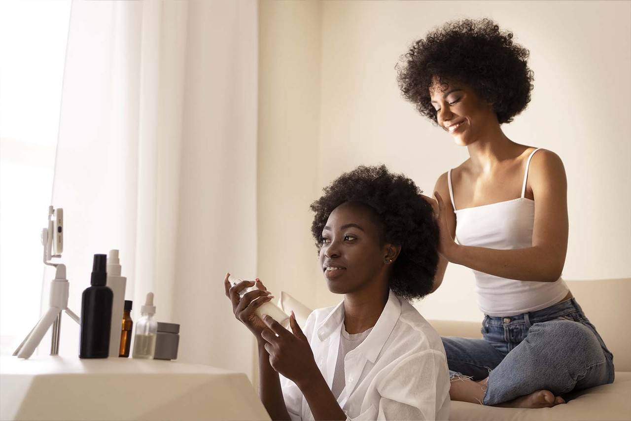 imagem de duas modelos de cabelos cacheados médios, em uma sala, uma segurando um produto para o cabelo, vestindo uma camiseta branca com uma camisa branca e outra modelo sentada logo atrás, com as mãos no cabelo da próxima, vestindo uma regata branca sorrindo