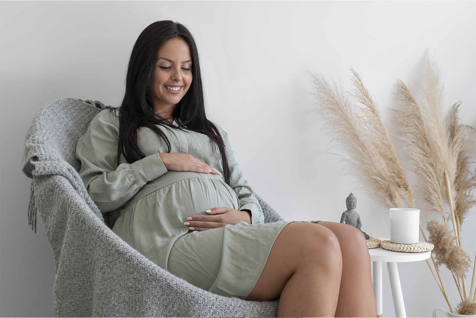 Imagem de uma mulher gravida vestindo um vestido verde bebe, sentada em um cadeira com as mãos na barriga em um fundo branco e cinza