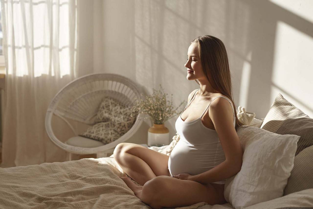 Imagem de uma mulher gravida de pijama sentada em sua cama de pernas cruzadas e as mãos na barriga de dia
